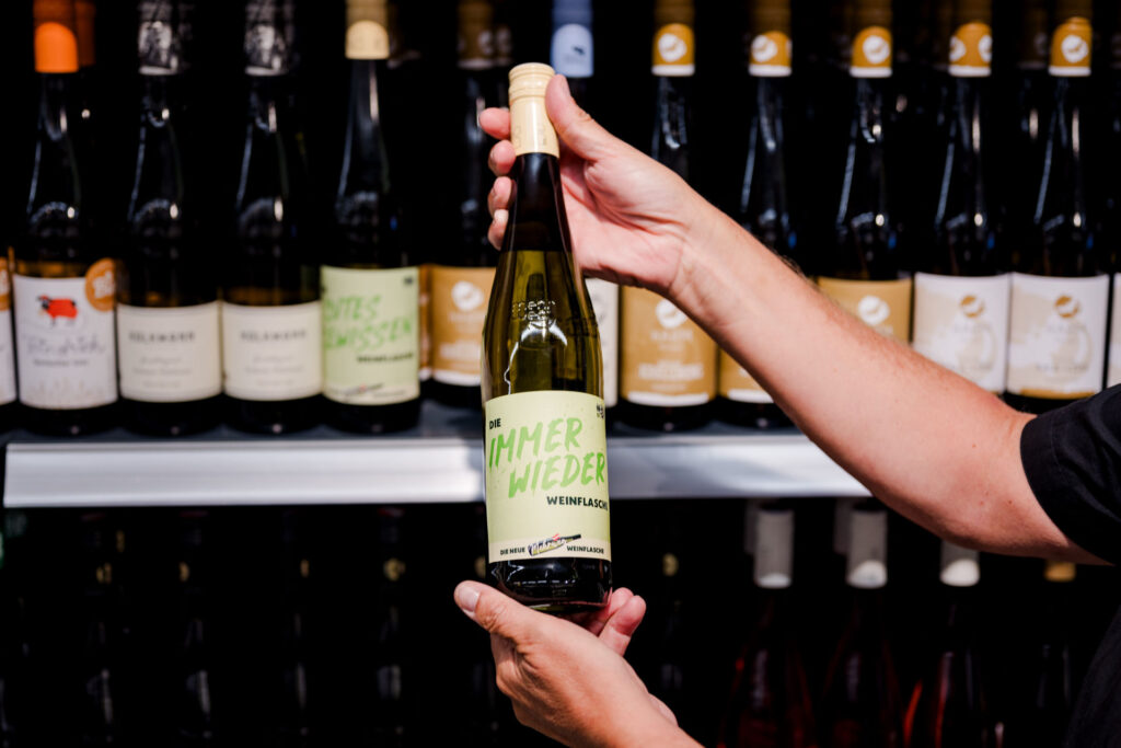 Die neue Mehrweg-Weinflasche – Immer wieder-Flasche vor Weinregal – zwei Hände