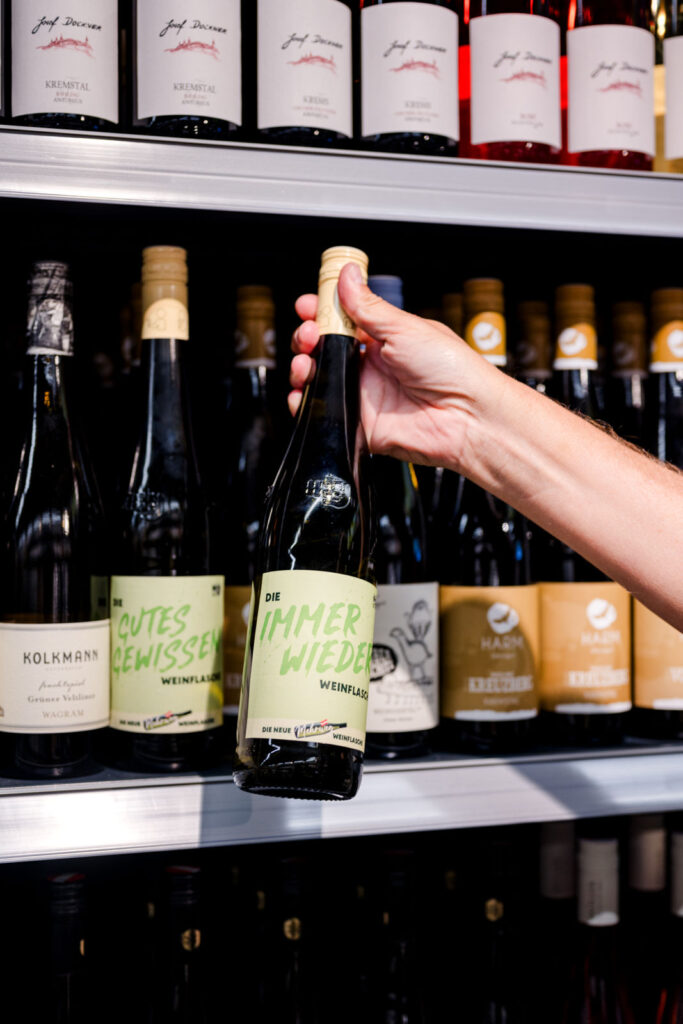 Die neue Mehrweg-Weinflasche – Immer wieder-Flasche vor Weinregal – eine Hand